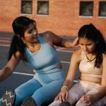 Mãe e filha fazendo alongamento juntas em uma pista esportiva ao ar livre.