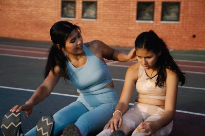 Mãe e filha fazendo alongamento juntas em uma pista esportiva ao ar livre.