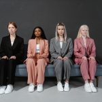 Quatro mulheres de diferentes perfis, vestidas com ternos elegantes, sentadas em um sofá em um estúdio