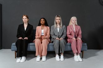 Quatro mulheres de diferentes perfis, vestidas com ternos elegantes, sentadas em um sofá em um estúdio